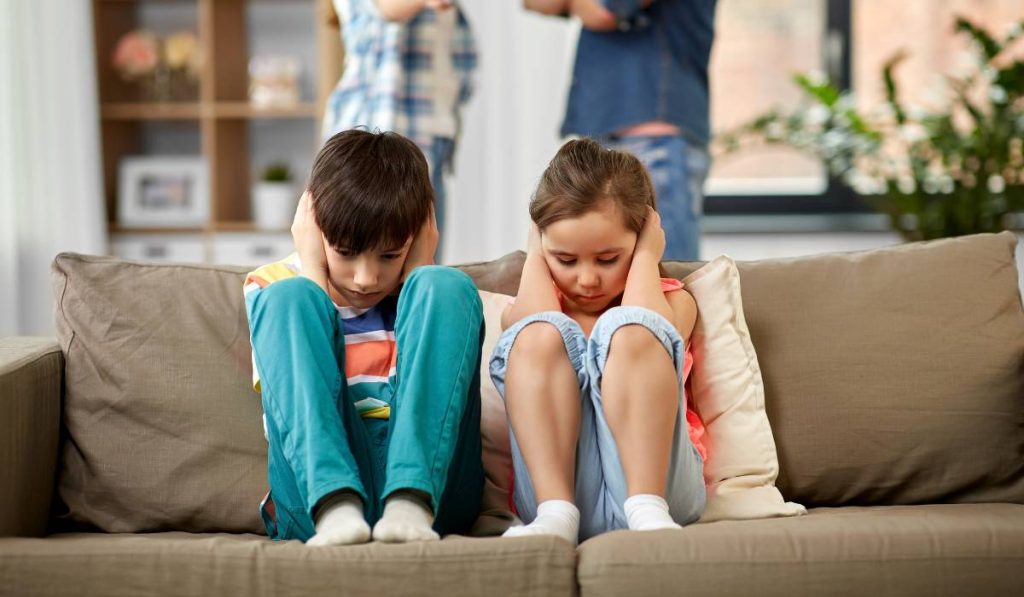 Two kids are sitting on a couch covering their ears with their hands while parents are having an arguement on the background.