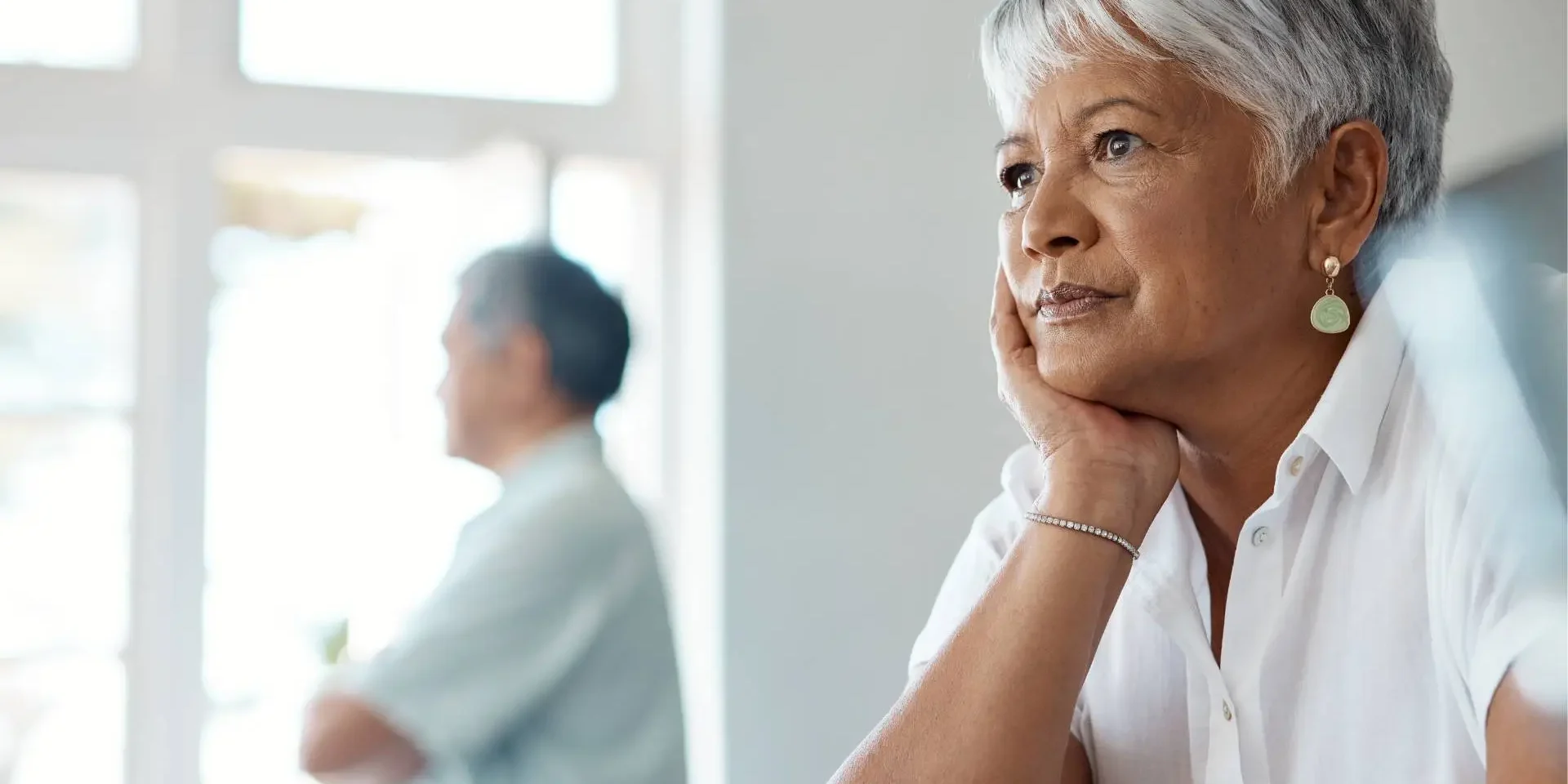 An old woman sitting at a table with a cup of coffee while her husband looking outside of the window with his hands folded.