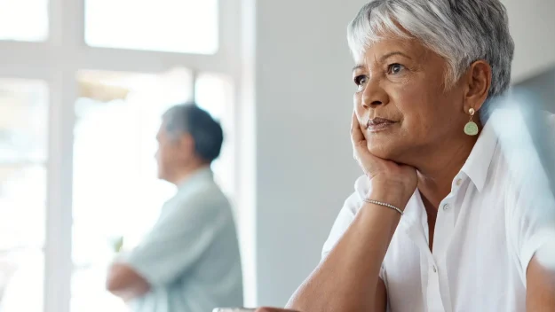 An old woman sitting at a table with a cup of coffee while her husband looking outside of the window with his hands folded.